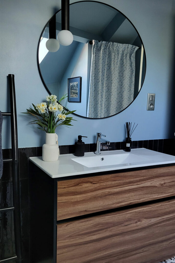 Salle de bain sous combles. Carrelage zellige noir en soubassement. Murs peints en bleu nuit. Grand miroir rond avec contour noir au dessus d'un meuble lavabo et une suspension lumineuse.