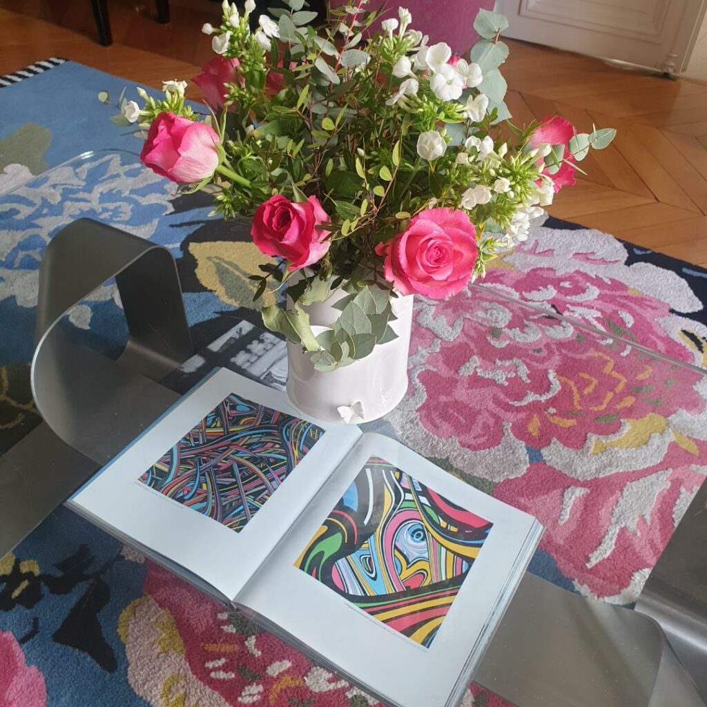 Bouquet de roses roses et fleurs blanches sur une table basse avec un livre d'art ouvert et un tapis floral coloré.