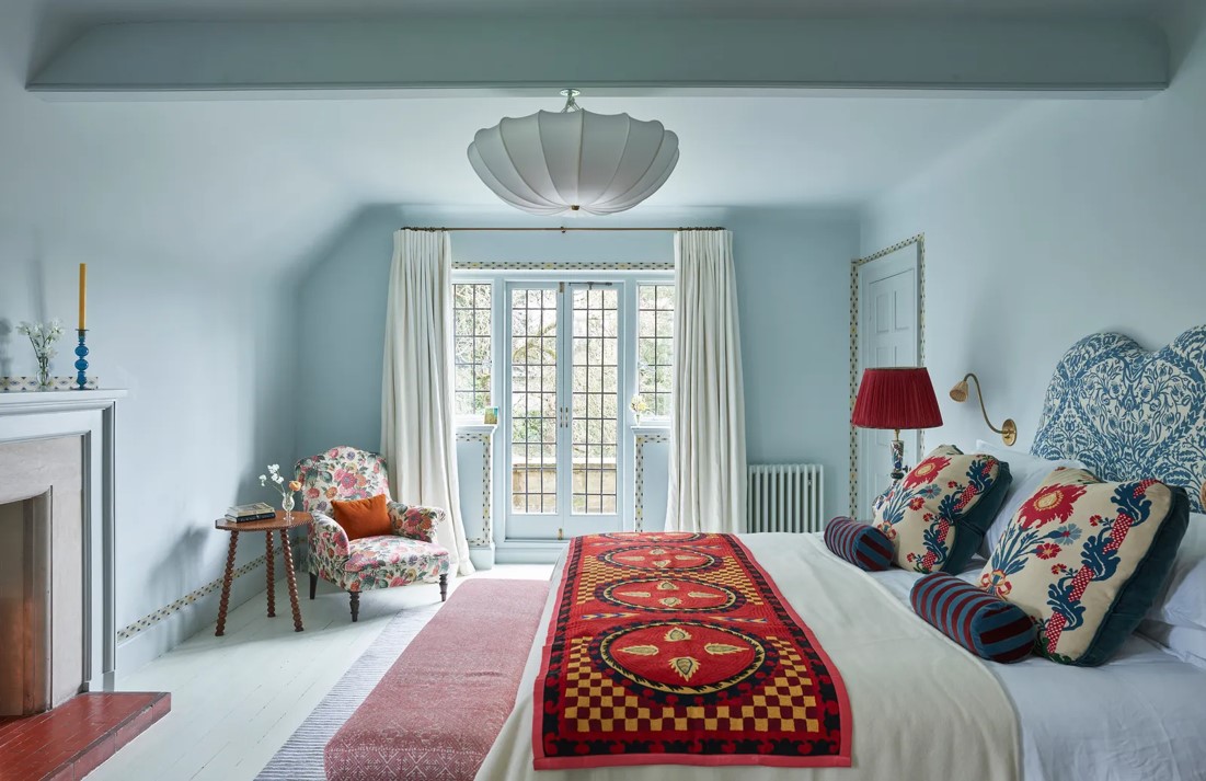 Chambre bleue style anglais avec rideaux blancs posés sol-plafond, cheminée et couvre-lit suzani floral rouge.