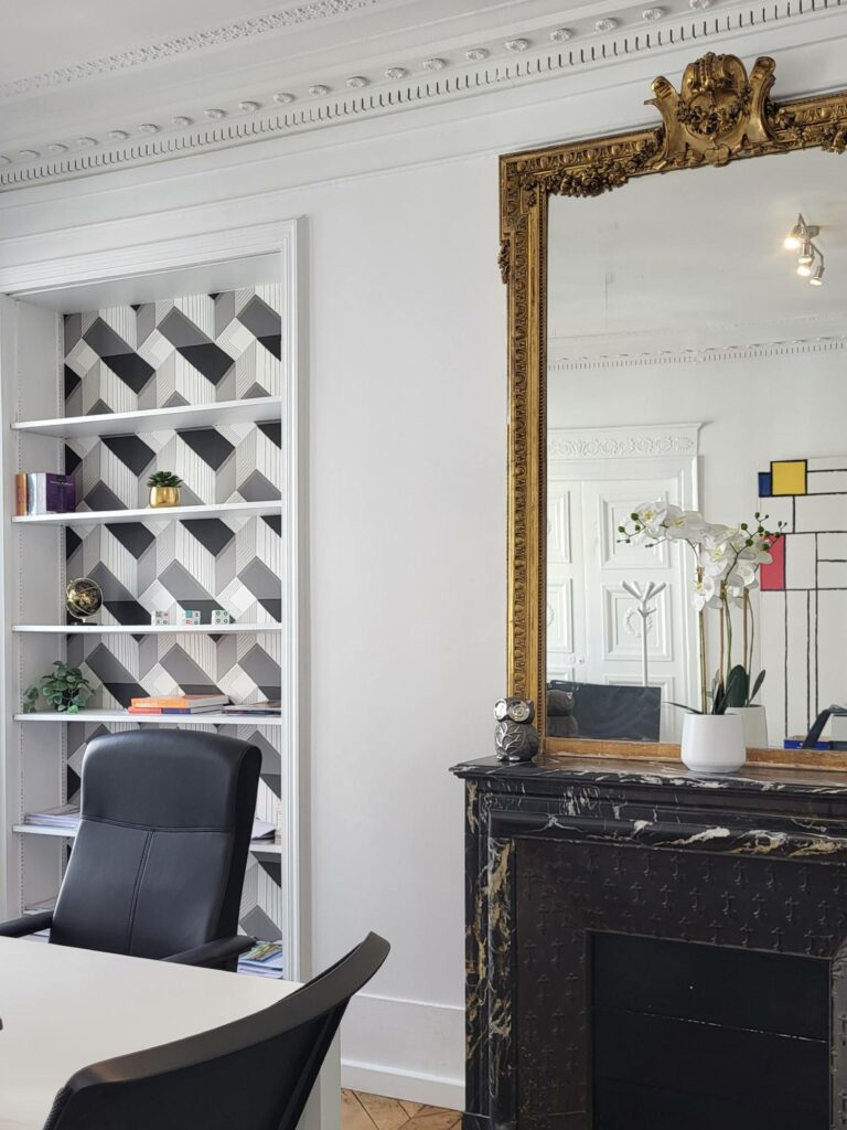Bureau avec bibliothèque encadrée habillée d'un papier peint géométrique noir et blanc, cheminée en marbre noir et miroir doré.