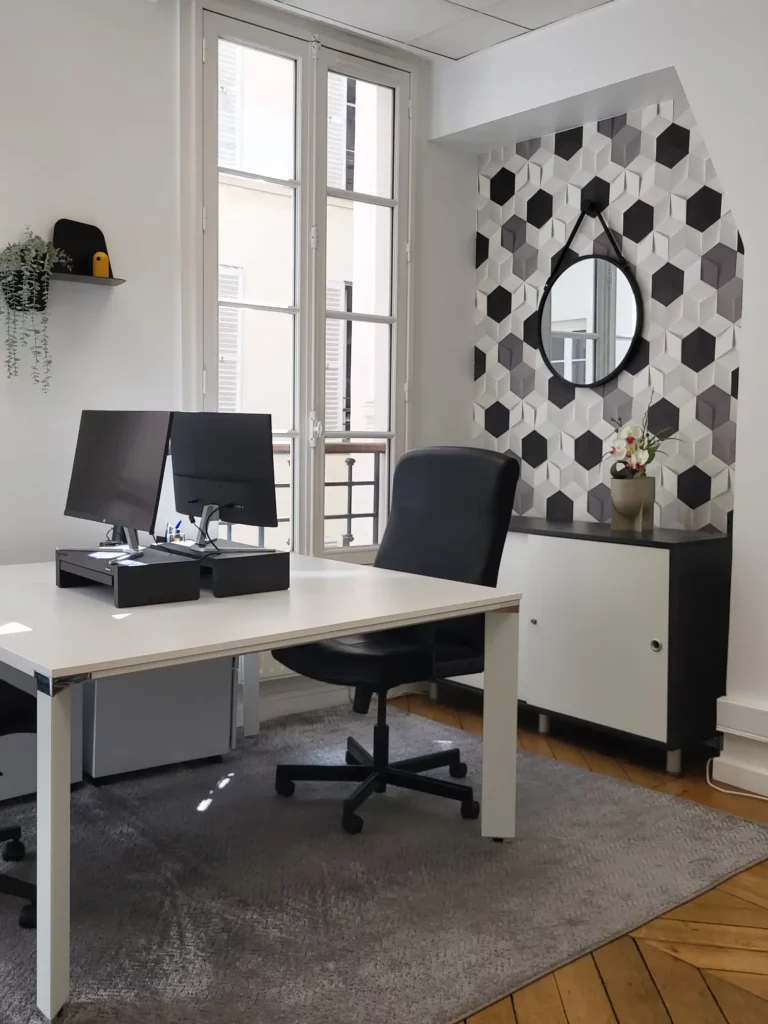 Bureau avec niche habillée de papier peint hexagonal noir, blanc et gris, miroir rond noir et meuble bas bicolore noir et blanc, deux bureaux individuels blancs avec ecrans.