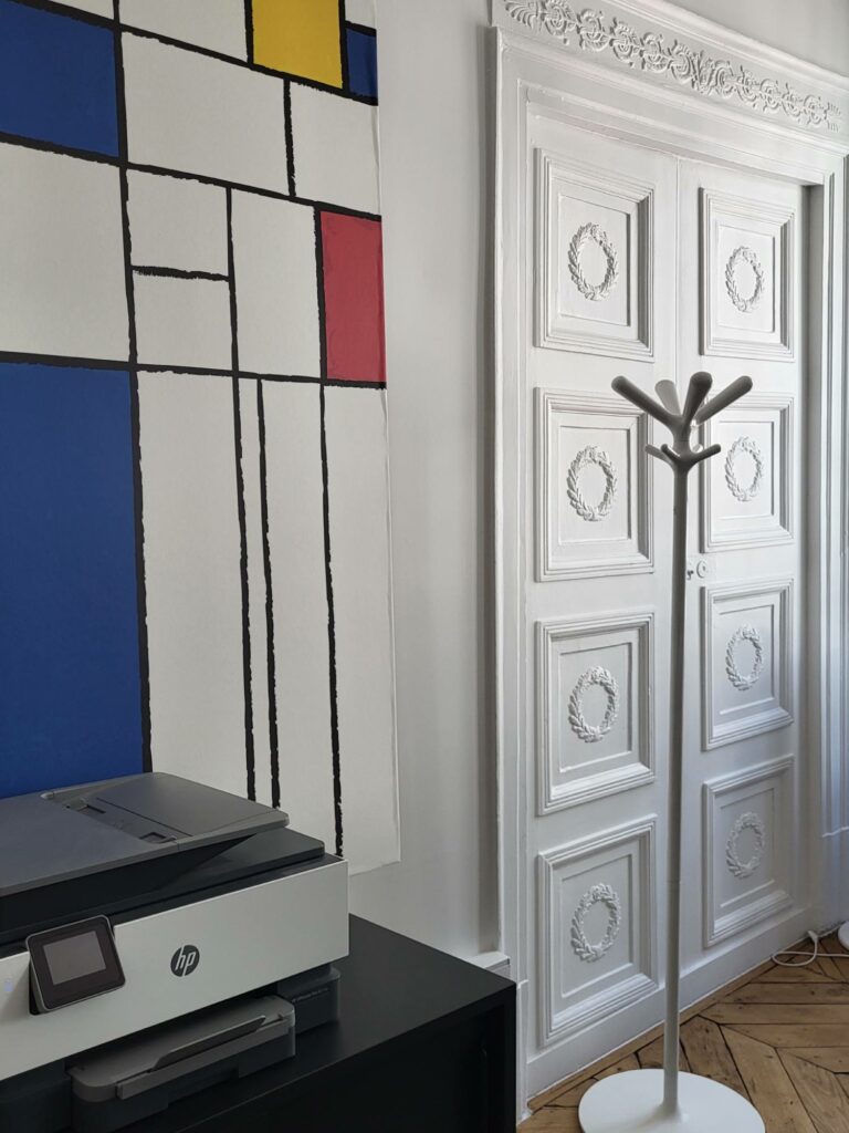 Bureau avec grand tableau inspiré Mondrian en bleu rouge jaune, porte à panneaux blancs haussmanniens et porte-manteau design blanc. Au premier plan - une imprimante de bureau.