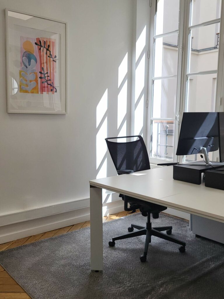 Bureau baigné de lumière naturelle avec grande fenêtre, œuvre d'art aquarelle colorée et bureau blanc avec chaises noires.