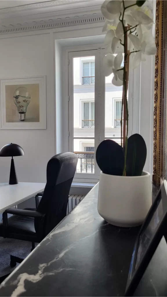 Bureau avec une lampe en métal noir et affiche représentant une ampoule-bocal avec des poissons. Cheminée en marbre noir et orchidée blanche.