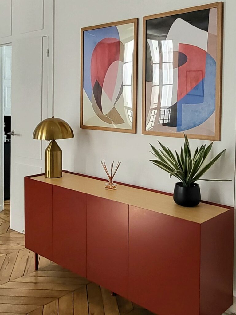 Entrée avec buffet laqué rouge terracotta et bois, lampe design métal dorée et deux tableaux abstraits encadrés chêne.