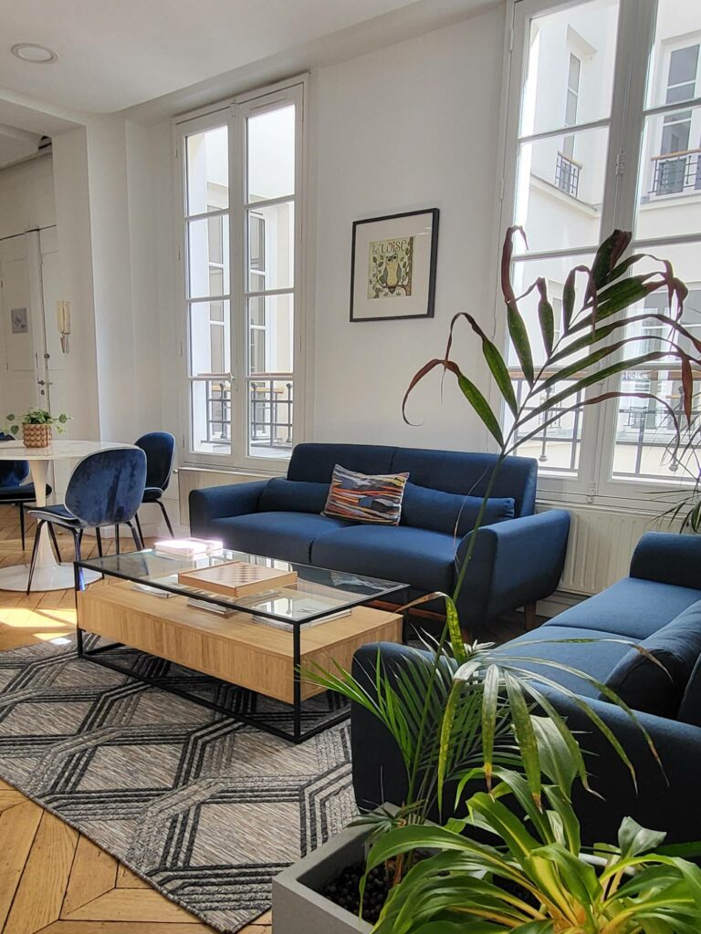 Espace avec deux canapés en tissu bleu et table basse en bois plateau en verre, espace avec table ronde plateau marbre blanc, chaises velours bleu marine, parquet chêne chevron.