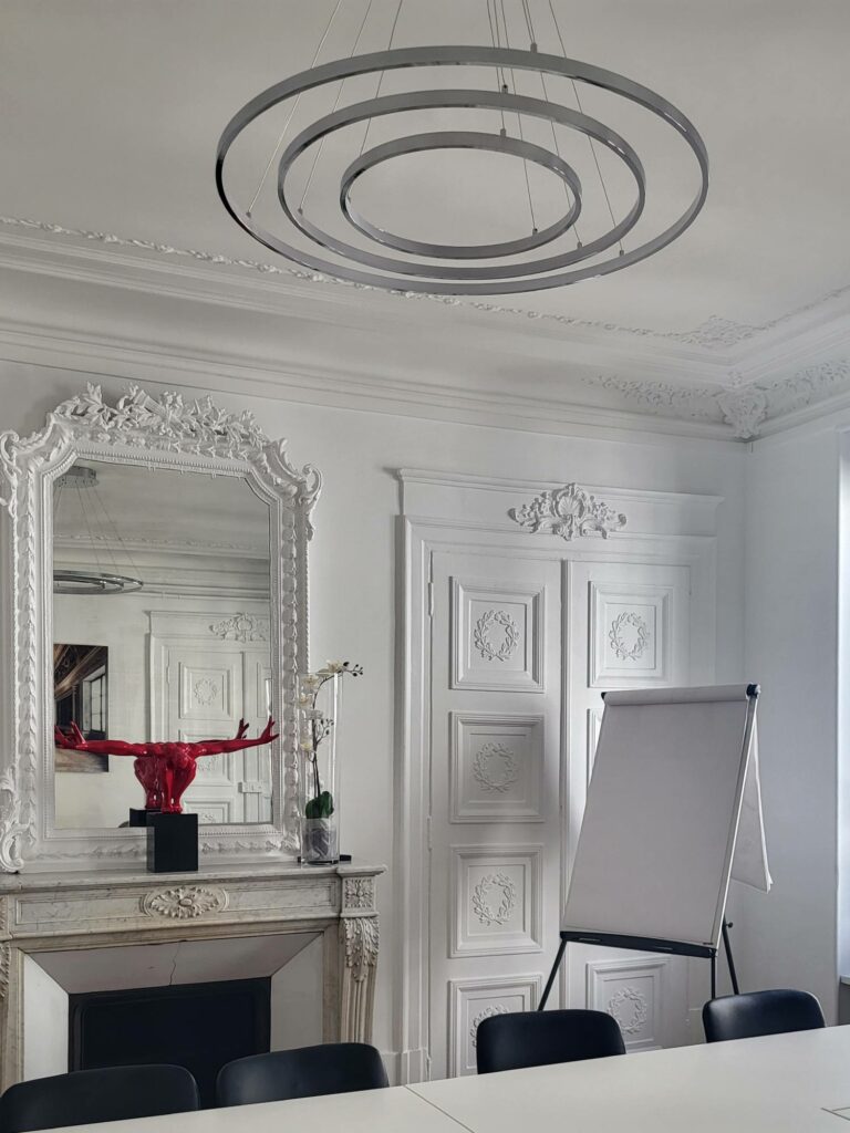 Salle de réunion avec cheminée baroque blanche, grand miroir sculpté blanc, suspension chromée circulaire, table de conférence et sculpture homme rouge sur la cheminée.