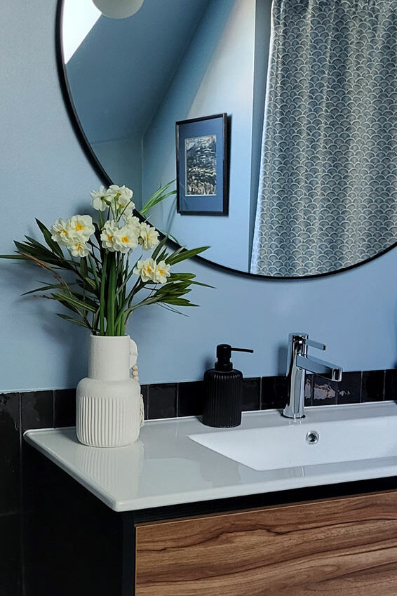 Salle de bain sous combles. Carrelage zellige noir en soubassement. Murs peints en bleu nuit. Grand miroir rond avec contour noir au dessus d'un meuble lavabo et une suspension lumineuse.